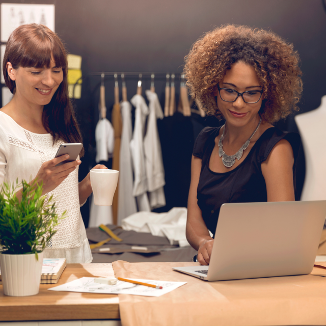 Deux femmes au comptoir d'un magasin de vêtements. L'une consulte son téléphone et l'autre son laptop au sujet de l'adaptation des statuts de société en Belgique (obligation légale à réaliser avant le 1er janvier 2024)